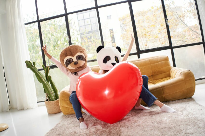 Two unrecognizable woman with animal mask bear panda holding big heart at home - Valentine's day people celebrating